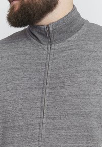 Maglione grigio con zip e colletto alto, tessuto testurizzato e cerniera nascosta. Il design presenta un aspetto morbido e informale.