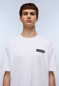 Hvid bomulds T-shirt med rund hals, korte ærmer og et sort rektangulært logo-patch med teksten "NAPAPIJRI" på venstre bryst.
