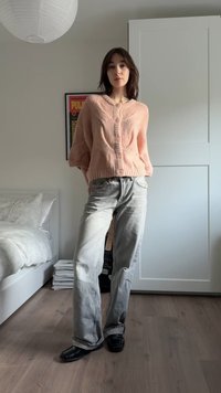 Ανοιχτό ροζ πλεκτή ζακέτα με υφασμένο σχέδιο, κουμπωμένη, συνδυασμένη με ανοιχτό γκρι τζιν με ίσια γραμμή και μαύρα παπούτσια τύπου slip-on.