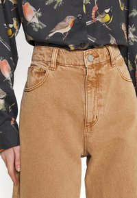 Bruna denim jeans med hög midja, med en slät yta, synliga sömmar och stängning fram med knapp, i kombination med en mönstrad skjorta med fåglar.