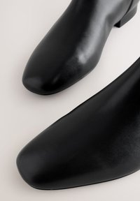 Paar schwarze Leder-Stiefeletten mit quadratischem und abgerundetem Zehendesign auf hellem Hintergrund.