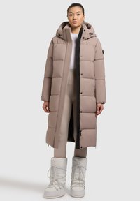 Haion lung, matlasat, de culoare taupe, cu glugă reglabilă prin șnur, fermoar frontal negru și buzunare laterale. Poartă peste pantaloni strâmți de culoare deschisă și cizme albe.