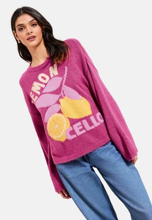 Femeie purtând un pulover roz moale cu un imprimeu de lămâi galbene și frunze mov, asortat cu blugi largi albaștri, pe un fundal alb.