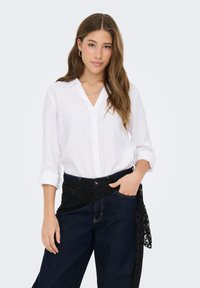 Chemise blanche à manches longues avec col et boutons sur le devant, associée à un pantalon en denim foncé avec une ceinture en dentelle noire.