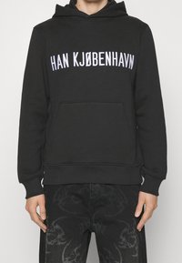 Felpa nera con tasca frontale, con la scritta bianca "HAN KJØBENHAVN". Realizzata in tessuto morbido con polsini e orlo a costine.