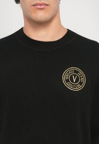 Μαύρο πλεκτό πουλόβερ με στρογγυλή λαιμόκοψη, που διαθέτει ένα χρυσό κεντημένο λογότυπο σε σχήμα κύκλου που γράφει "Versace Jeans Couture".