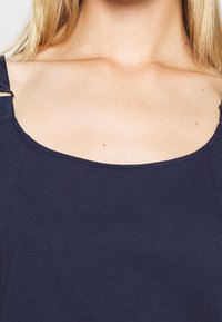 Close-up van een persoon met een lichte huid, die een marineblauwe mouwloze top draagt met een vierkante halslijn en verstelbare schouderbandjes.