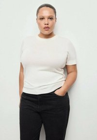 Kremasta pletena majica s kratkimi rokavi in okrolim izrezom, kombinirana s črnimi kavbojkami. Tkanina je videti gladka, z oprijeto silhueto.