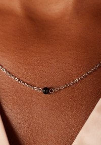 Zilveren ketting met een centrale zwarte kraal, geplaatst tegen een gladde, warm getinte huid met een zachte stof in de hoek.