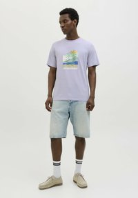 Camiseta de manga corta color lavanda con estampado gráfico, combinada con pantalones cortos de denim azul claro, calcetines blancos con rayas negras y zapatos beige.