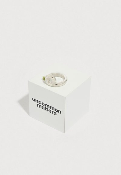 Bague en argent avec un dessus circulaire et une touche verte, présentée sur une boîte carrée blanche avec l'inscription "uncommon matters" en noir.