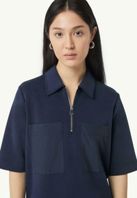 Marineblauwe polo met korte mouwen, een ritskraag en twee voorzakken, met een gladde textuur en een ontspannen pasvorm.