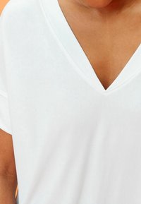 Zbliżenie na osobę ubraną w gładką, białą koszulkę z dekoltem w serek i krótkim rękawem na pomarańczowym tle.