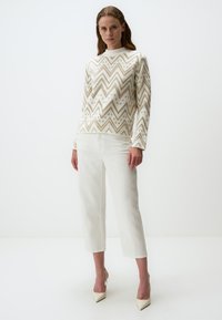 Witte gebreide trui met een gouden zigzagpatroon, hoge hals en lange mouwen, gecombineerd met witte cropped broek en lichtgekleurde hakken.