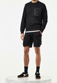 Homme portant un sweat-shirt noir à manches longues avec une poche zippée à l'avant, un short cargo noir, des chaussettes noires et des baskets noires, debout sur une plateforme en marbre.