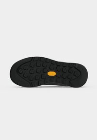 Semelle de chaussure noire avec des crampons en forme de croix et un logo Vibram orange au centre sur un fond blanc uni.