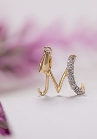 Gouden hanger in de vorm van de letter "M," met aan één kant kleine, heldere edelstenen die langs de kromming zijn ingebed.