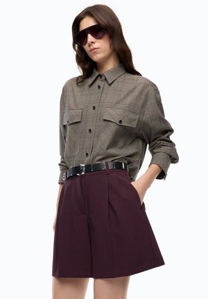 Donna con grandi occhiali da sole scuri, camicia grigia a quadri con tasche, cintura nera e pantaloncini bordeaux a vita alta con le mani nelle tasche.