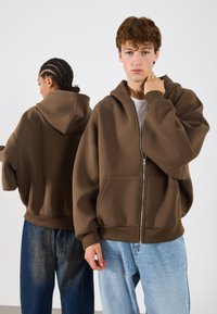 Rjava zip-up kapuca s prostornim krojem, velikimi sprednjimi žepi, rebrastimi manšetami in čezmerno kapuco, v kombinaciji z svetlo modrimi densami.