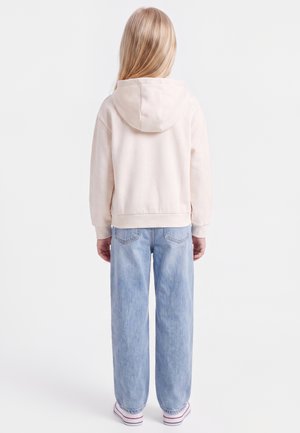 Enfant aux longs cheveux blonds portant un sweat à capuche beige clair, un jean bleu clair et des baskets blanches, dos tourné sur un fond uni.
