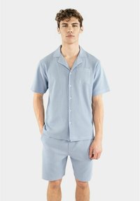 Vaaleansininen pyjama-setti, joka koostuu lyhythihaisesta kauluspaidasta ja polvimittaisista shortseista. Valmistettu teksturoidusta materiaalista, jossa on pystysuorat raidat.