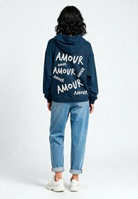 Felpa navy con testo bianco "AMOUR" di diverse dimensioni sulla schiena, abbinata a jeans azzurri arrotolati e sneakers bianche.