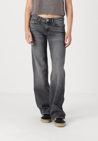 Žena v obnošených čiernych bootcut džínsoch, čiernych teniskách s béžovými podrážkami a skrátenom šedom topánku, stojí na pozadí čistej bielej steny.