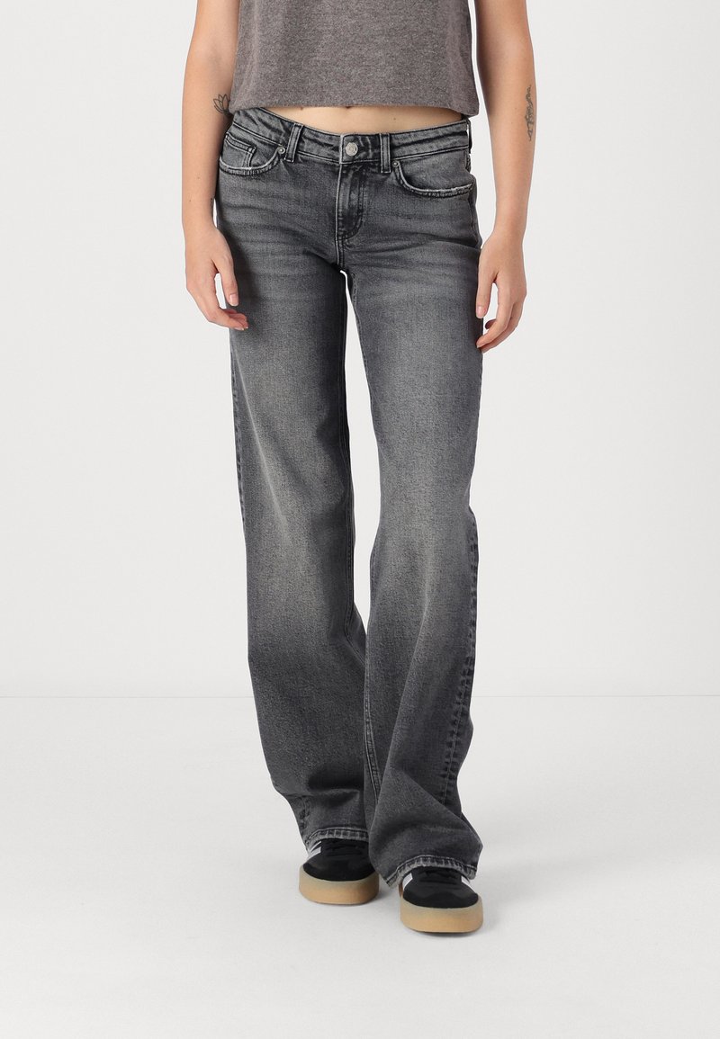 Žena v obnošených čiernych bootcut džínsoch, čiernych teniskách s béžovými podrážkami a skrátenom šedom topánku, stojí na pozadí čistej bielej steny.