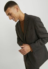 Blazer in tweed scuro con un sottile motivo a macchie, taglio sartoriale, rever a scollo e taschino sul petto. Indossato sopra un maglione marrone.