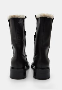 Botas de tobillo de cuero negro con un cuello forrado de piel de oveja, que presentan una textura suave, cremalleras laterales y un tacón grueso negro.
