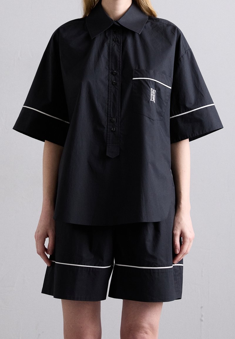 Personne portant une chemise noire ample à manches courtes boutonnée et un short assorti avec une bordure blanche et un petit logo brodé sur la poche de la chemise.