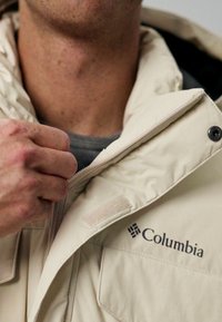 Een hand van een persoon die de rits van een lichtbeige Columbia jacket omhoog trekt, met het merkembleem op het borstgebied.