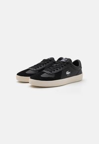 Zapatillas negras con parte superior de ante y cuero, diseño con cordones y suela de goma en color crema contrastante. Se puede ver el logo de Lacoste.