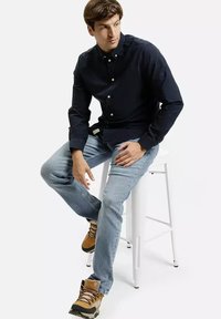 Ciemnoniebieska koszula zapinana na guziki, jasne dżinsy i jasnobrązowe buty trekkingowe. Koszula ma kołnierzyk i guziki, podczas gdy dżinsy są nieco postrzępione.