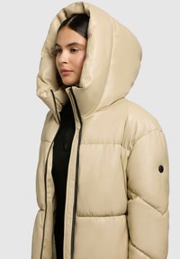 Bežová prešívaná bunda s nadrozmernou kapucňou, zipsovým uzáverom a čiernymi akcentami. Má prešívanú textúru a pod ňou je čierny rebrovaný top.