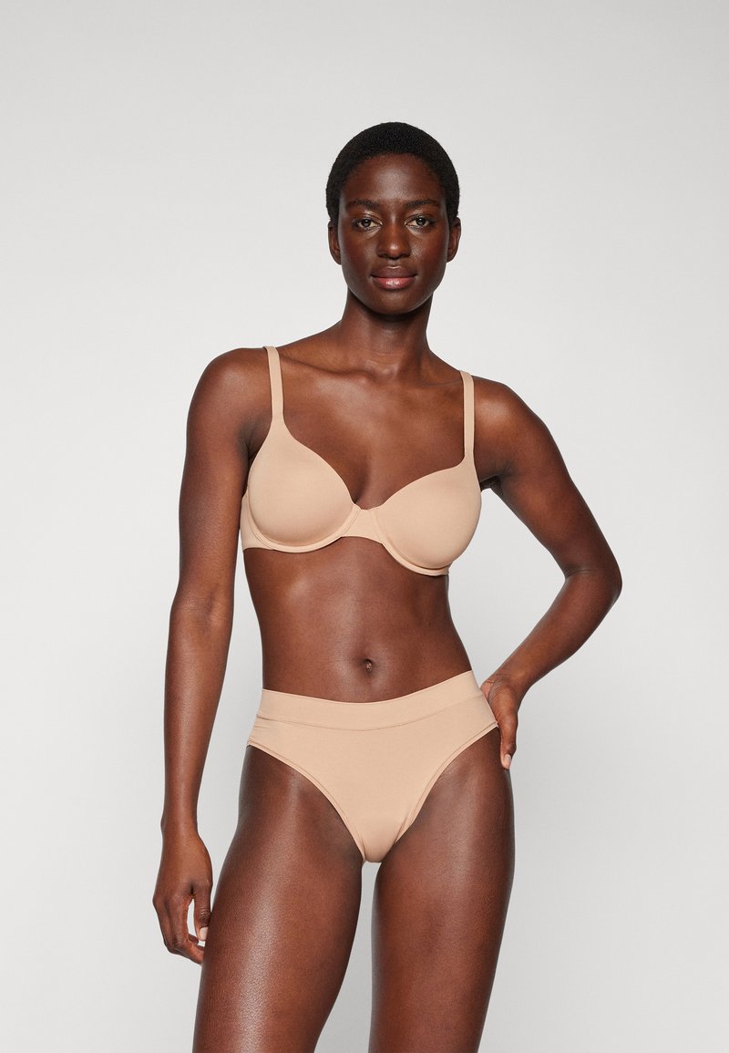 Beige beha met gevormde cups en verstelbare straps, gecombineerd met bijpassende hoge bikini slips. Glad, rekbaar materiaal, naadloos ontwerp.