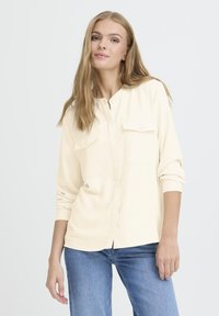 Light beige button-up shirt with two chest pockets, long sleeves, and a relaxed fit, paired with blue denim jeans.