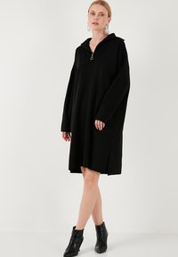 Rochie neagră supradimensionată, cu guler cu fermoar până la un sfert, mâneci lungi, slituri pe laterale și o textură moale. Purtați-o cu bocanci negri strălucitori, până la glezne.