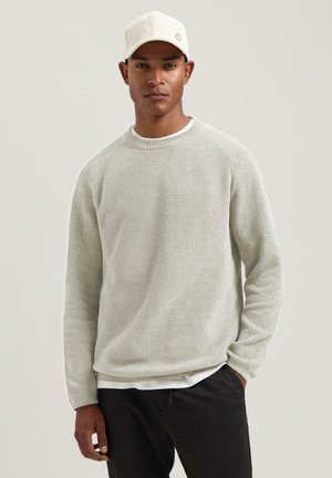 Lichtgrijze gebreide trui met een ronde hals, gedragen over een wit shirt, gecombineerd met donkere broek en aangevuld met een witte pet.