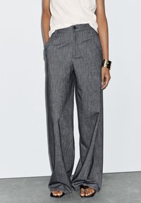 Pantalons larges gris sur mesure avec une texture subtile, poches avant, fermeture à bouton et taille haute, associés à un haut léger.