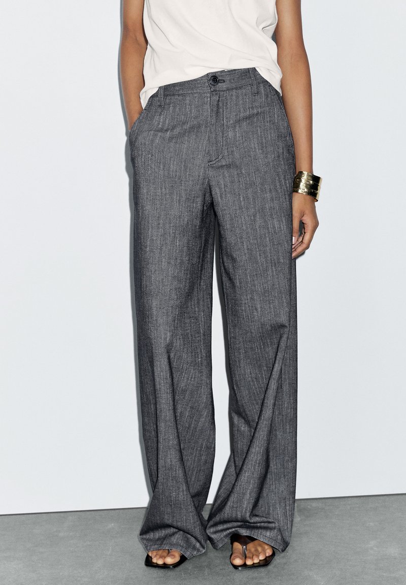 Pantalons larges gris sur mesure avec une texture subtile, poches avant, fermeture à bouton et taille haute, associés à un haut léger.