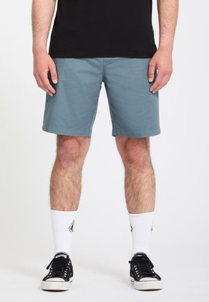 Man draagt blauwe shorts, witte sokken tot halverwege de kuit met logo, zwarte lage sneakers en een zwart T-shirt, staand op een witte achtergrond.