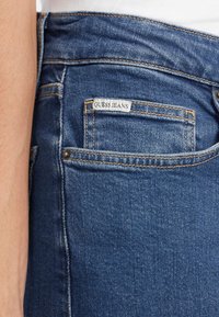 Plave traperice s izblijedjelim završetkom, koje na stražnjoj strani imaju malu bijelu oznaku s natpisom "GUESS JEANS" i kontrastne šavove.