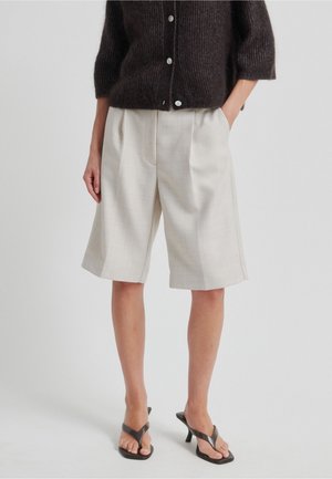 Beige skræddersyede shorts med plisseringer, lavet af tekstureret stof. Kombineret med en mørk, strikket, cropped sweater og sorte klip-klappere.