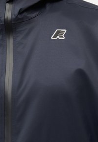 Σκούρο μπλε αδιάβροχο μπουφάν με μαύρο αδιάβροχο φερμουάρ και λευκό-μαύρο λογότυπο στο στήθος.