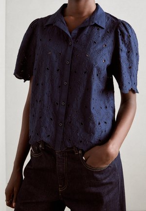 Woman wearing navy blue embroidered short-sleeve button-up blouse with scalloped edges and dark denim jeans, one hand in pocket.