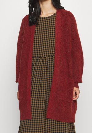 Femme portant un long cardigan rouge en tricot ample par-dessus une robe à carreaux marron et noir, la main reposant près d'une grande poche avant.