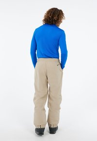 Blauwe langemouwen shirt gecombineerd met beige losse broek. De broek heeft een elastische tailleband en verstelbare manchetten. Zwarte schoenen zichtbaar.