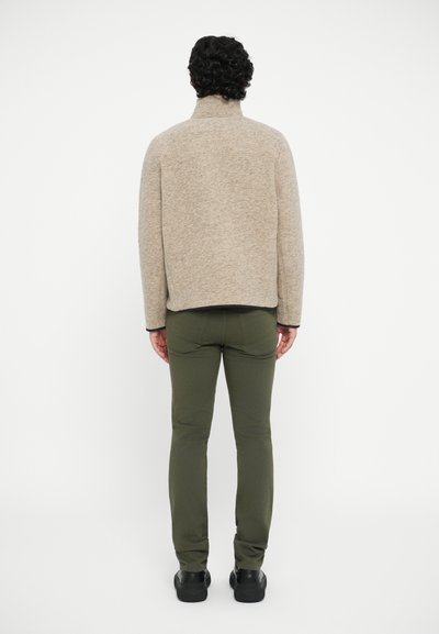 Beige, textured high-collared jacket, dark trim, paired with olive green pants and black shoes, viewed from the back.