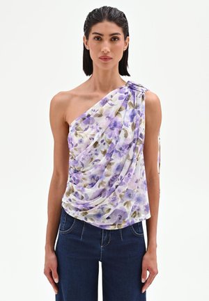 Femme portant un top blanc à une épaule avec imprimé floral violet et un jean bleu, debout devant un fond clair uni.